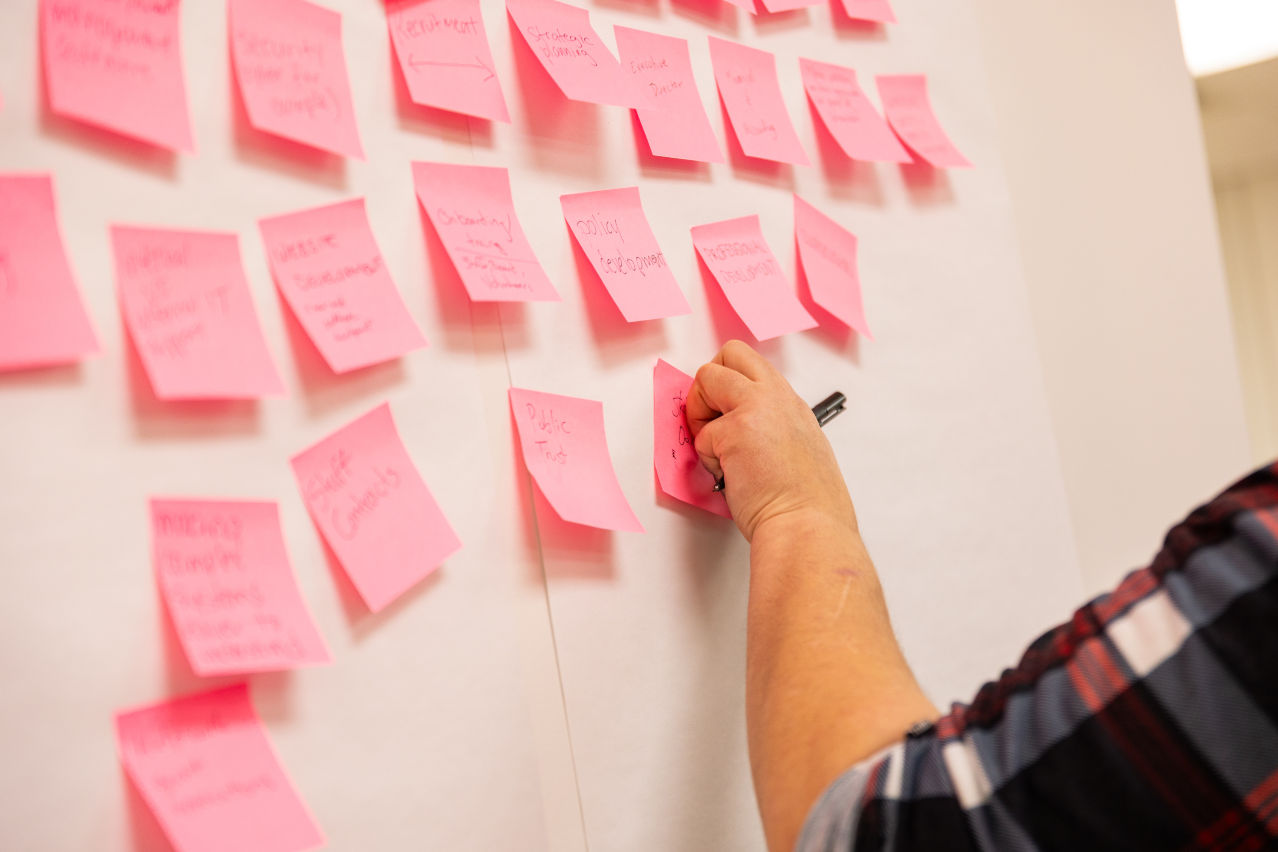 An arm reaches to add a pink sticky note with writing on it to a board with over 15 additional sticky notes.