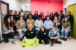 20-25 people, a mix of seated and standing, posing for a group photo with a bright pink background at the end of a long day of connecting.