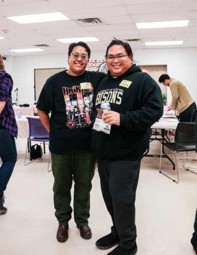 Two people smile and posing for a photo with arms around each other. The one on the left is in dark green pants and a graphic t-shirt and the one on the right is dressed in black pants and a black University of Manitoba hoodie.